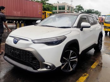 A white compact crossover SUV with sporty alloy wheels and a modern front grille, parked on a wet pavement. The vehicle features a blue emblem on the grille and is positioned near a shipping container yard, with a person walking nearby.