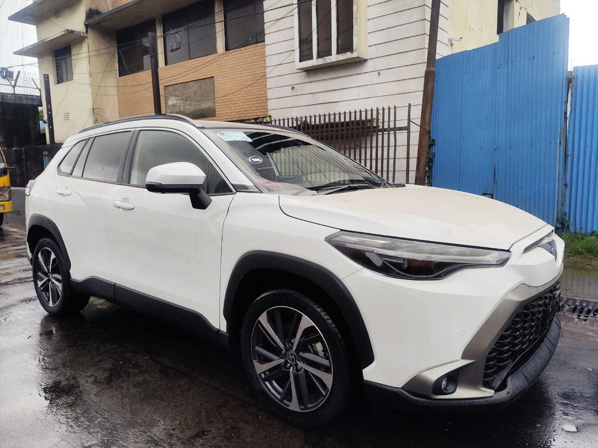 Car Planet A white compact crossover SUV with sporty alloy wheels and a modern front grille, parked on a wet pavement. The vehicle features a blue emblem on the grille and is positioned near a shipping container yard, with a person walking nearby.