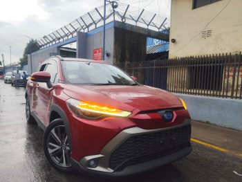 A red 2021 Toyota Corolla Cross Z Package Hybrid is parked on a wet road, likely in front of a gated industrial area. The car features a sleek design with prominent LED headlights and black alloy wheels. The Toyota emblem on the front grille is visible, and the car's indicators are on, emitting a bright amber glow. The background shows a fence with barbed wire and a few people walking in the distance, with overcast skies suggesting recent rainfall