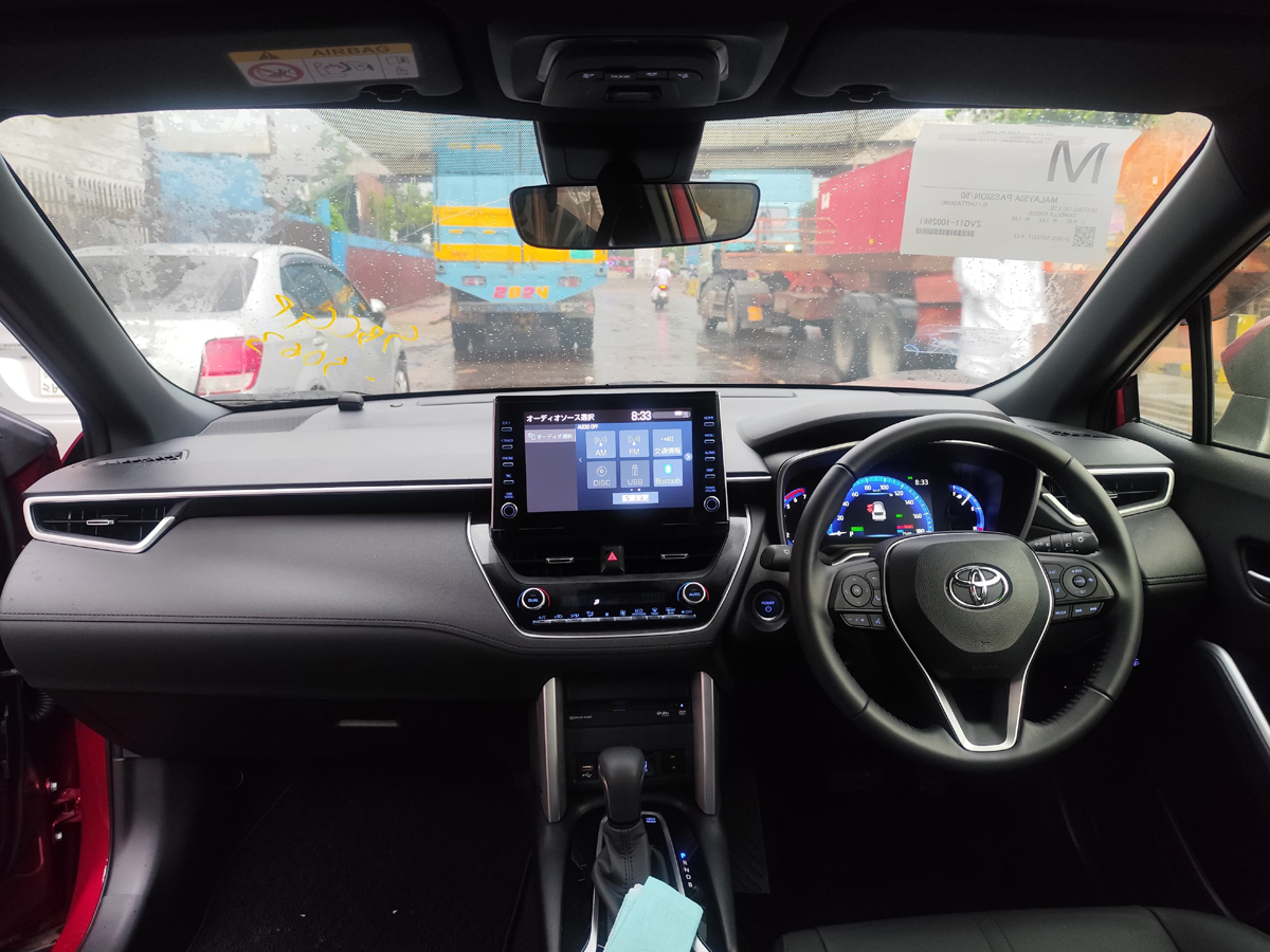 Car Planet A red 2021 Toyota Corolla Cross Z Package Hybrid is parked on a wet road, likely in front of a gated industrial area. The car features a sleek design with prominent LED headlights and black alloy wheels. The Toyota emblem on the front grille is visible, and the car's indicators are on, emitting a bright amber glow. The background shows a fence with barbed wire and a few people walking in the distance, with overcast skies suggesting recent rainfall