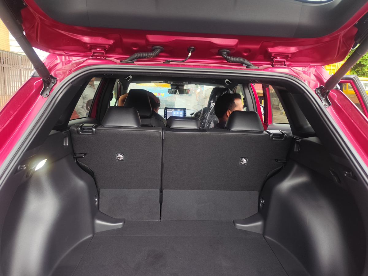 Car Planet A red 2021 Toyota Corolla Cross Z Package Hybrid is parked on a wet road, likely in front of a gated industrial area. The car features a sleek design with prominent LED headlights and black alloy wheels. The Toyota emblem on the front grille is visible, and the car's indicators are on, emitting a bright amber glow. The background shows a fence with barbed wire and a few people walking in the distance, with overcast skies suggesting recent rainfall