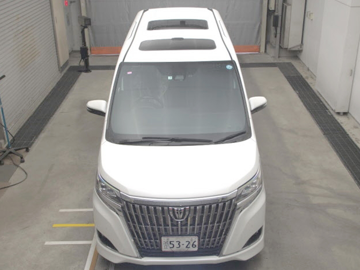 Car Planet A luxurious 2019 Toyota Esquire Gi minivan in pristine white is parked indoors. The vehicle features a sleek and modern design, with a prominent chrome grille and sharp, sophisticated headlights. The minivan's clean lines and spacious body are accentuated by large windows and stylish alloy wheels, giving it an elegant and upscale appearance. The license plate and a small "JU" logo are visible, indicating it is from a Japanese auction.