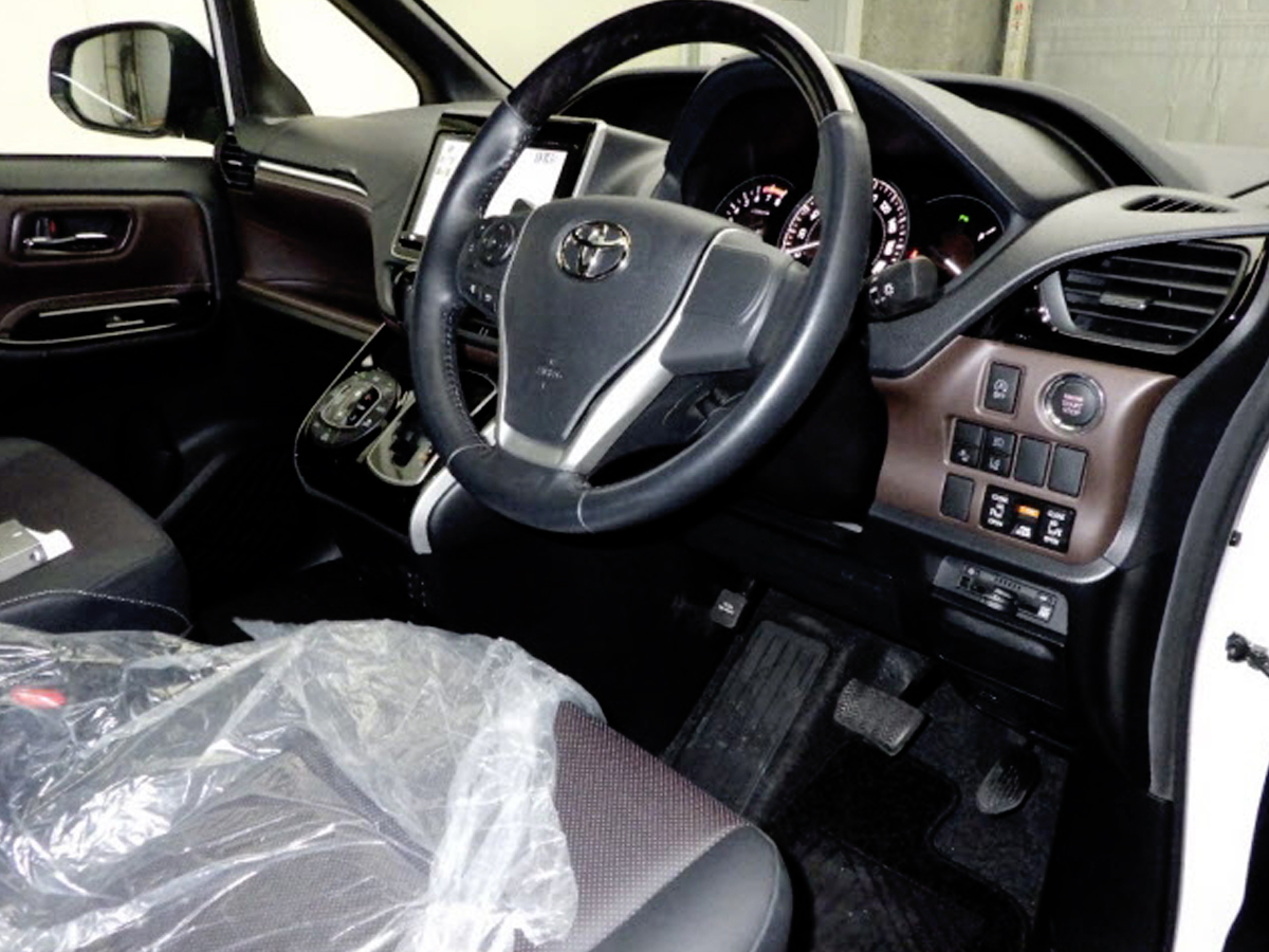 Car Planet A luxurious 2019 Toyota Esquire Gi minivan in pristine white is parked indoors. The vehicle features a sleek and modern design, with a prominent chrome grille and sharp, sophisticated headlights. The minivan's clean lines and spacious body are accentuated by large windows and stylish alloy wheels, giving it an elegant and upscale appearance. The license plate and a small "JU" logo are visible, indicating it is from a Japanese auction.