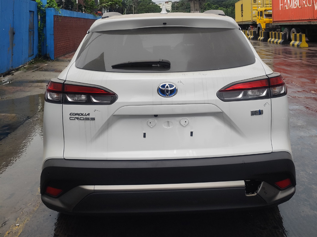 Car-Planet A white compact crossover SUV with sporty alloy wheels and a modern front grille, parked on a wet pavement. The vehicle features a blue emblem on the grille and is positioned near a shipping container yard, with a person walking nearby.