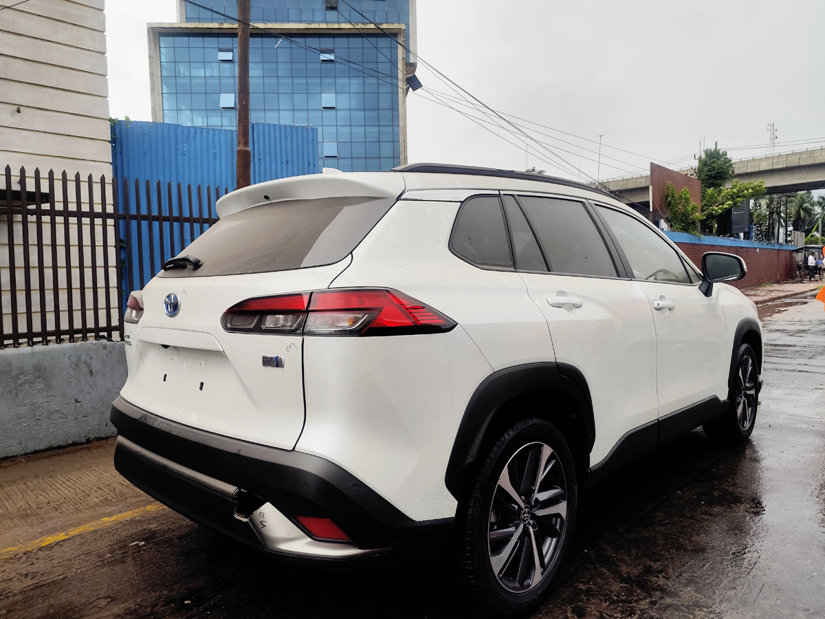 Car-Planet A white compact crossover SUV with sporty alloy wheels and a modern front grille, parked on a wet pavement. The vehicle features a blue emblem on the grille and is positioned near a shipping container yard, with a person walking nearby.