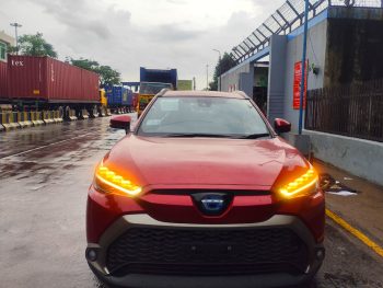 A red 2021 Toyota Corolla Cross Z Package Hybrid is parked on a wet road, likely in front of a gated industrial area. The car features a sleek design with prominent LED headlights and black alloy wheels. The Toyota emblem on the front grille is visible, and the car's indicators are on, emitting a bright amber glow. The background shows a fence with barbed wire and a few people walking in the distance, with overcast skies suggesting recent rainfall