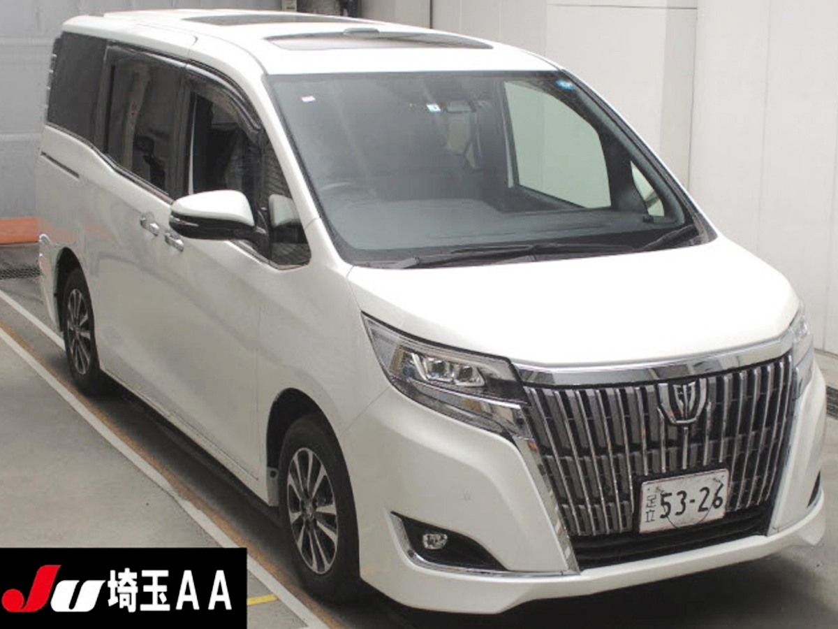 Car Planet A luxurious 2019 Toyota Esquire Gi minivan in pristine white is parked indoors. The vehicle features a sleek and modern design, with a prominent chrome grille and sharp, sophisticated headlights. The minivan's clean lines and spacious body are accentuated by large windows and stylish alloy wheels, giving it an elegant and upscale appearance. The license plate and a small "JU" logo are visible, indicating it is from a Japanese auction.
