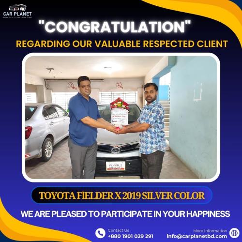 A congratulatory banner from Car Planet featuring two men shaking hands while holding a flower bouquet in front of a Toyota Fielder X 2019 silver car. The banner reads Congratulations to a respected client on the purchase of the vehicle. The text below states Toyota Fielder X 2019 Silver Color and We are pleased to participate in your happiness. The company’s contact details are provided at the bottom.