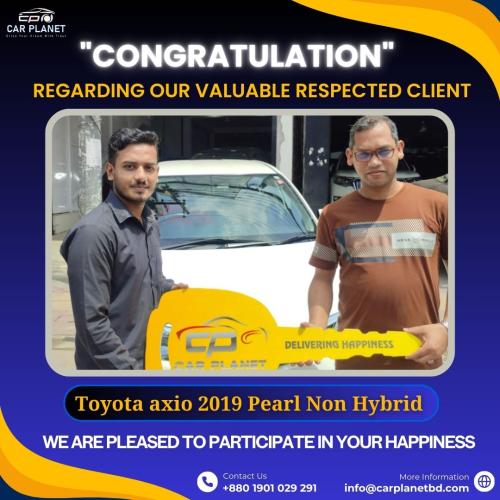 Car Planet representative handing over a key-shaped plaque to a satisfied customer in front of a white Toyota Axio 2019 Pearl Non-Hybrid car, celebrating the purchase with a congratulatory message. Contact information for Car Planet is also displayed.