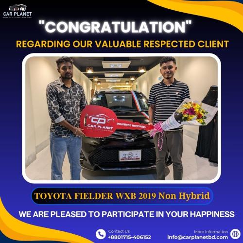The image shows a happy customer receiving a black Toyota Fielder WXB 2019 Non-Hybrid at Car Planet dealership. One person is holding a symbolic key with the Car Planet logo, while the other holds a bouquet of flowers, celebrating the purchase. The dealership background features a well-lit showroom. The banner congratulates the valued customer, emphasizing Car Planet's commitment to Delivering Happiness. Contact details and additional information about buying cars in Bangladesh are displayed, making this an ideal promotional image for car sales and customer satisfaction.