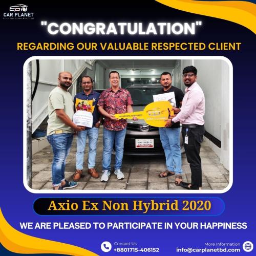 The image shows a celebratory moment where five men are standing in front of a Toyota Axio Ex Non-Hybrid 2020 vehicle, smiling and holding a large symbolic key. The backdrop reads CONGRATULATION REGARDING OUR VALUABLE RESPECTED CLIENT. One of the men is holding a bouquet of flowers, while another holds the oversized key that says Delivering Happiness with the logo of Car Planet. The text below reads, Axio Ex Non Hybrid 2020. We are pleased to participate in your happiness. Contact information and the company logo are displayed at the bottom.